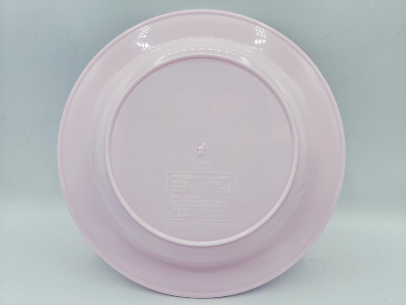 Puede incluir: Un plato de pl&aacute;stico rosa claro con un borde ligeramente elevado. El plato tiene una superficie lisa y un dise&ntilde;o circular. La parte inferior del plato tiene un peque&ntilde;o centro elevado con texto y s&iacute;mbolos en relieve.