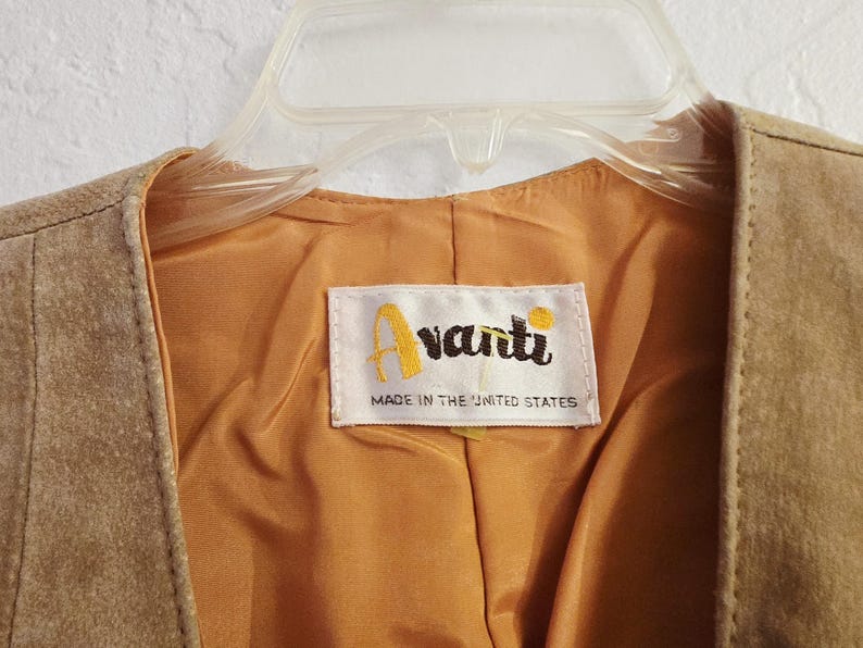 May include: Close-up of a tan suede vest with a gold satin lining. The vest is hanging on a clear plastic hanger. A label inside the vest reads "Avanti" and "Made in the United States".