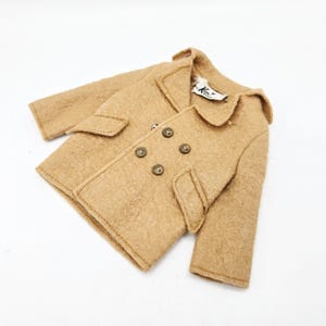 May include: A tan wool coat with a double-breasted design, featuring four dark brown buttons and two front pockets. The coat has a classic collar and is likely for a child.