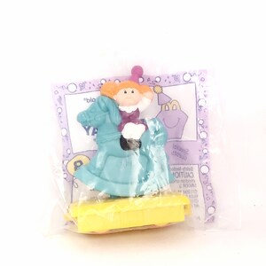 May include: A plastic toy set featuring a girl riding a rocking horse. The girl has red hair and is wearing a purple dress. The rocking horse is blue and yellow. The toy is packaged in a clear plastic bag with a purple and white background.