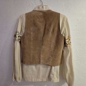 May include: A tan suede vest layered over a cream-colored blouse with embroidered sleeves. The vest has a tailored fit, and the blouse features a collared neckline and long sleeves. The sleeves have decorative embroidery.