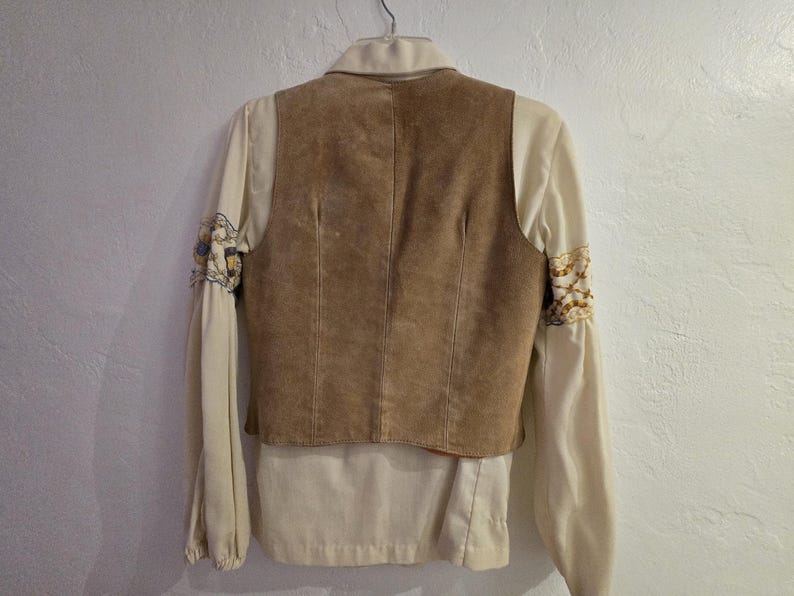 May include: A tan suede vest layered over a cream-colored blouse with embroidered sleeves. The vest has a tailored fit, and the blouse features a collared neckline and long sleeves. The sleeves have decorative embroidery.