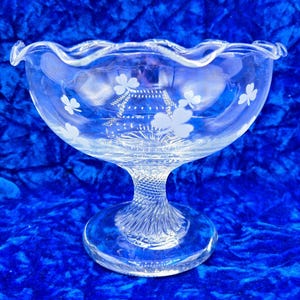 May include: A clear glass pedestal bowl with a scalloped rim and etched shamrock designs. The bowl is on a blue velvet surface.
