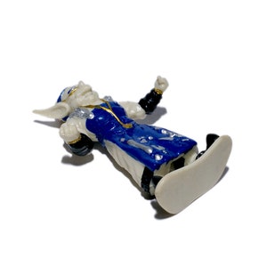 Puede incluir: Una figurita de pl&aacute;stico blanca y azul de un goblin que lleva una t&uacute;nica azul y dorada, montando una tabla de snowboard blanca. El goblin tiene una cara blanca con ojos negros y una boca negra.