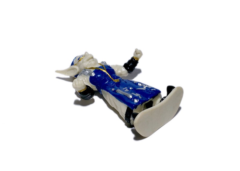 Puede incluir: Una figurita de pl&aacute;stico blanca y azul de un goblin que lleva una t&uacute;nica azul y dorada, montando una tabla de snowboard blanca. El goblin tiene una cara blanca con ojos negros y una boca negra.