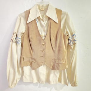 May include: A vintage cream-colored blouse with long sleeves and embroidered details on the cuffs. A tan suede vest with a button-up front is layered over the blouse. The outfit is hanging on a hanger against a white background.
