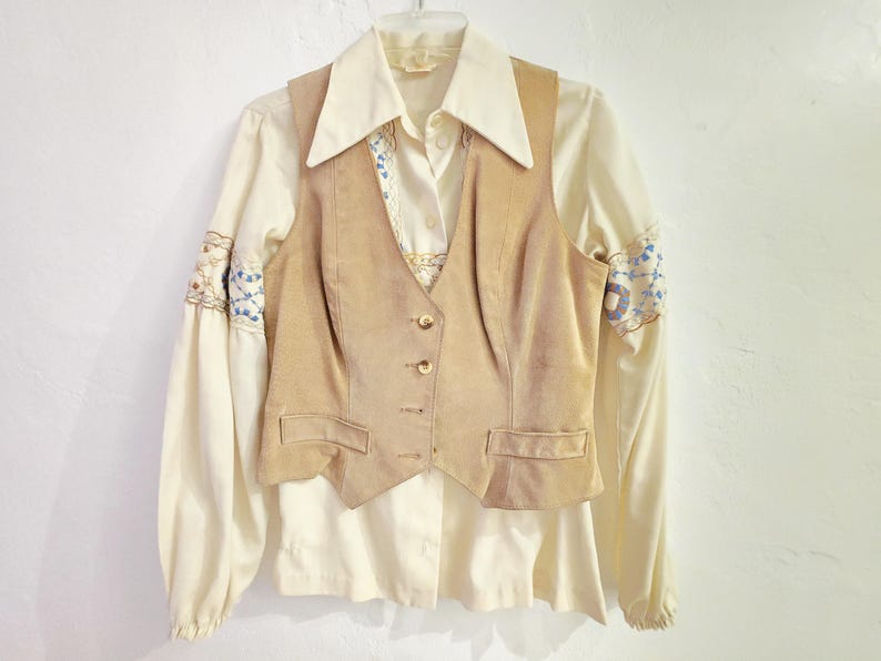 May include: A vintage cream-colored blouse with long sleeves and embroidered details on the cuffs. A tan suede vest with a button-up front is layered over the blouse. The outfit is hanging on a hanger against a white background.