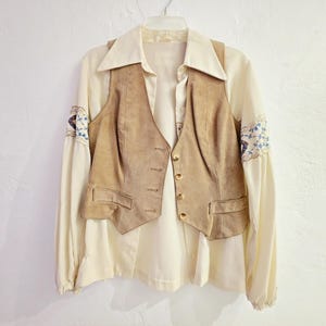 May include: Cream-colored blouse with a tan suede vest. The blouse has long sleeves with blue and white embroidered details on the cuffs. The vest has a button-up front and two pockets. The outfit is hanging on a white hanger.