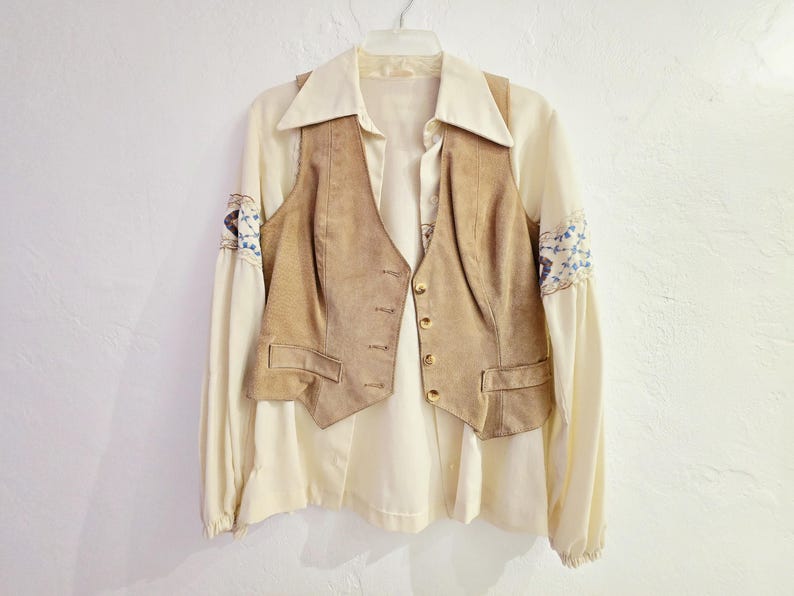May include: Cream-colored blouse with a tan suede vest. The blouse has long sleeves with blue and white embroidered details on the cuffs. The vest has a button-up front and two pockets. The outfit is hanging on a white hanger.