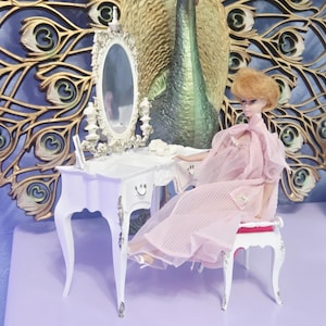 Op de afbeelding: Een pop in een roze jurk zit aan een witte kaptafel met een spiegel. De kaptafel heeft een lachend gezichtsontwerp. Een decoratieve wandophanging met pauwenthema staat op de achtergrond. De pop draagt een roze jurk en zit op een witte stoel met een rode zitting.