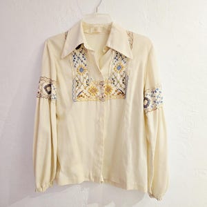 May include: Cream-colored blouse with long sleeves and a collared neckline. The blouse features intricate embroidery in blue, yellow, and brown on the front and sleeves. The embroidery has a floral design.