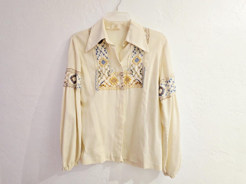 May include: Cream-colored blouse with long sleeves and a collared neckline. The blouse features intricate embroidery in blue, yellow, and brown on the front and sleeves. The embroidery has a floral design.