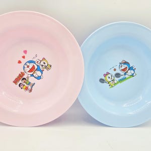 Puede incluir: Dos platos de pl&aacute;stico, uno rosa y otro azul claro, cada uno con un dibujo animado. El plato rosa tiene un dise&ntilde;o de Doraemon con corazones, mientras que el plato azul muestra a Doraemon jugando al tenis. Ambos platos tienen un borde ancho.