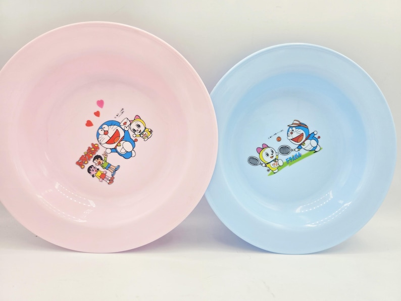 Puede incluir: Dos platos de pl&aacute;stico, uno rosa y otro azul claro, cada uno con un dibujo animado. El plato rosa tiene un dise&ntilde;o de Doraemon con corazones, mientras que el plato azul muestra a Doraemon jugando al tenis. Ambos platos tienen un borde ancho.