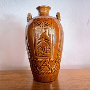 Puede incluir: Un jarrón de cerámica marrón con cuello estrecho y dos asas pequeñas. El jarrón presenta caracteres chinos en relieve y detalles en forma de cuerda. El jarrón mide aproximadamente 25 cm de alto.