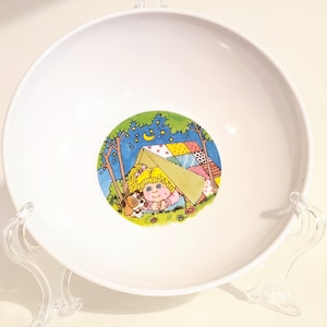 Può includere: Una ciotola in ceramica bianca con un'illustrazione colorata di una ragazza e il suo cane all'interno di una tenda. L'illustrazione è circondata da un cerchio verde e lo sfondo è blu con stelle.