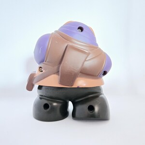 May include: A purple and brown plastic toy figure with a black base. The figure has a purple head and a brown body with a strap across the chest. The figure is standing on a white surface.