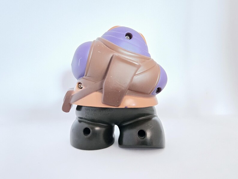 May include: A purple and brown plastic toy figure with a black base. The figure has a purple head and a brown body with a strap across the chest. The figure is standing on a white surface.