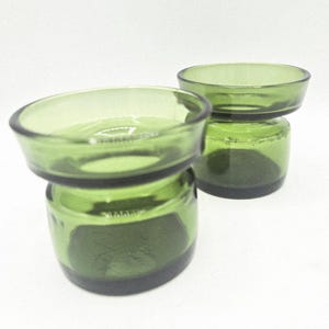 Op de afbeelding: Twee groene glazen kandelaars. Elk heeft een kleine, ronde komvorm bovenop een cilindrische basis. Het glas heeft een lichte groene tint en het algehele ontwerp is eenvoudig en elegant. De kandelaars staan tegen een witte achtergrond.