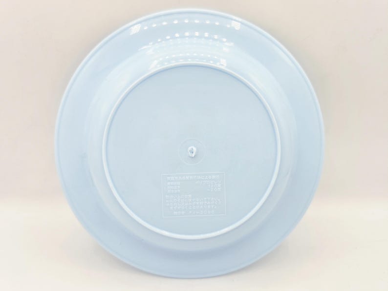 Puede incluir: Un plato de pl&aacute;stico azul claro con un borde ligeramente elevado. El plato tiene un dise&ntilde;o circular con una hendidura central. La parte posterior del plato tiene una peque&ntilde;a etiqueta rectangular con texto japon&eacute;s.