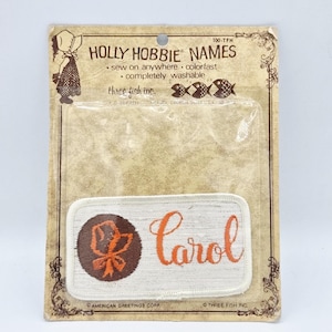 May include: A vintage Holly Hobbie name patch with the name "Carol" in orange script. The patch features a brown and orange floral design on a white background. The packaging includes the text "Holly Hobbie Names" and "sew on anywhere".