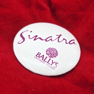 以下が含まれることがあります： 白い丸いピンバッジ。紫色の筆記体で「Sinatra」と書かれています。下には、紫色のBally's Las Vegas Nevadaのロゴがあります。赤いテクスチャの背景にピンバッジが置かれています。