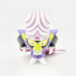 May include: A Mojo Jojo figurine featuring a large white brain-shaped head with purple accents, a lime green face with yellow eyes, a purple collar, black arms, white gloves, and a purple cape. The character has a stern expression with visible teeth.
