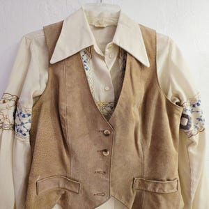 May include: A vintage ensemble featuring a tan suede vest with button closures and two front pockets. Underneath, a cream-colored blouse with embroidered sleeves and a collared neckline is visible. The outfit is hung on a white hanger.