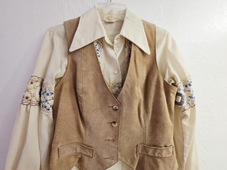 May include: A vintage ensemble featuring a tan suede vest with button closures and two front pockets. Underneath, a cream-colored blouse with embroidered sleeves and a collared neckline is visible. The outfit is hung on a white hanger.
