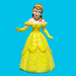 May include: A yellow figurine of a princess in a ball gown, with matching gloves and a headband. The figure has brown hair and is set against a bright blue background. The figurine is a collectible item.