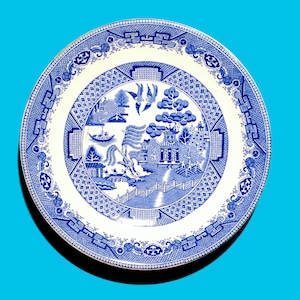May include: A blue and white porcelain plate with a traditional Chinese willow pattern design. The plate features a central image of a pagoda, a willow tree, and a bridge.