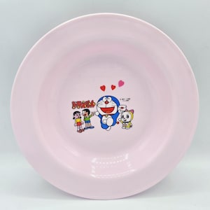 Puede incluir: Un cuenco redondo de cer&aacute;mica rosa claro con una ilustraci&oacute;n de dibujos animados de Doraemon y sus amigos. La imagen incluye los personajes Doraemon, Nobita, Shizuka y Dorami, con corazones encima. El cuenco tiene un borde ancho.