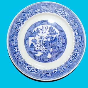 May include: A white ceramic plate with a blue and white willow pattern. The plate features a traditional Chinese landscape scene with a bridge, trees, and a pagoda.