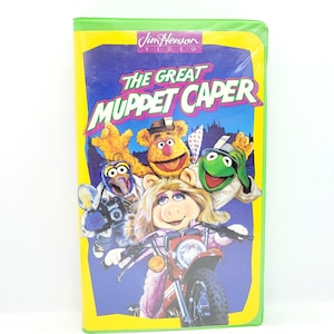 May include: A VHS tape featuring The Great Muppet Caper. The cover features Miss Piggy riding a motorcycle with Kermit the Frog, Fozzie Bear, and Gonzo in the background. The tape is green and yellow with the title in bold, black letters.