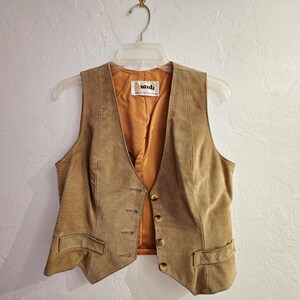 May include: A tan suede vest with a V-neck and button closure. The vest has two front pockets and a gold-colored interior lining. The vest is hanging on a clear plastic hanger against a white wall.