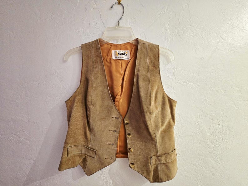 May include: A tan suede vest with a V-neck and button closure. The vest has two front pockets and a gold-colored interior lining. The vest is hanging on a clear plastic hanger against a white wall.