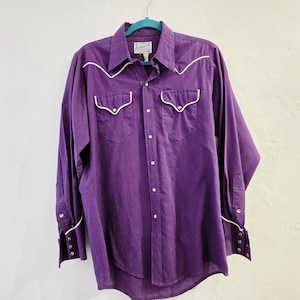 Op de afbeelding: Een paars Western-shirt met lange mouwen en witte biezen langs de kraag, zakken en manchetten. Het shirt heeft twee voorzakken en witte knopen. Het shirt hangt aan een turquoise hanger.