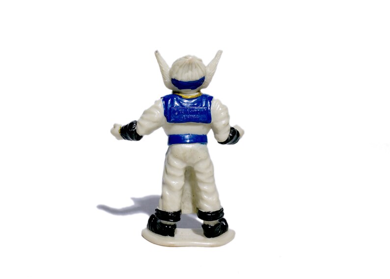 Puede incluir: Una figura de acci&oacute;n de pl&aacute;stico blanco con detalles azules y dorados. La figura tiene un casco blanco con una visera azul y una pieza de pecho azul y dorada. La figura est&aacute; de pie sobre una base blanca.