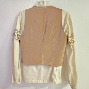 May include: Back view of a vintage cream-colored blouse with a tan suede vest. The long sleeves have embroidered details in gold, blue, and cream. The vest has vertical seams and a collared neckline.