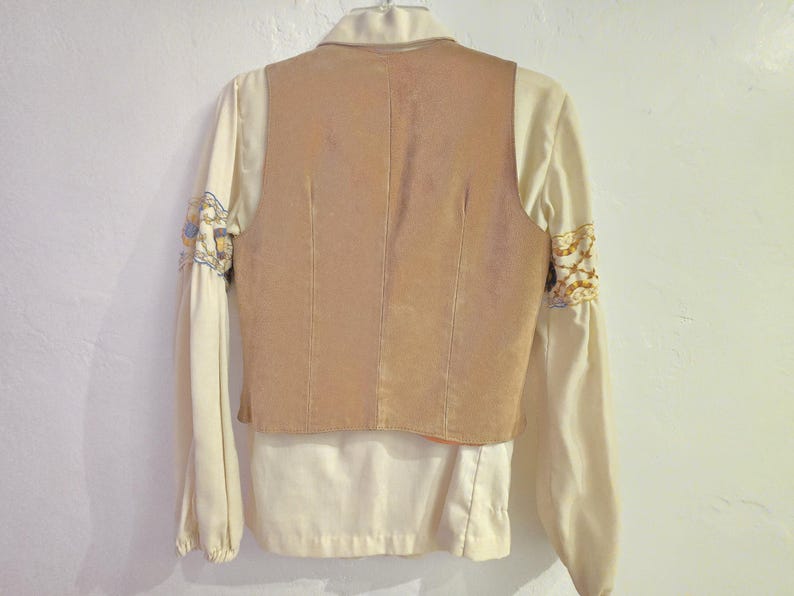 May include: Back view of a vintage cream-colored blouse with a tan suede vest. The long sleeves have embroidered details in gold, blue, and cream. The vest has vertical seams and a collared neckline.
