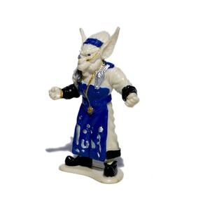 Puede incluir: Una figurita de pl&aacute;stico blanca de una criatura fant&aacute;stica con detalles azules y dorados. La criatura tiene orejas grandes, una nariz puntiaguda y lleva una t&uacute;nica azul y blanca.