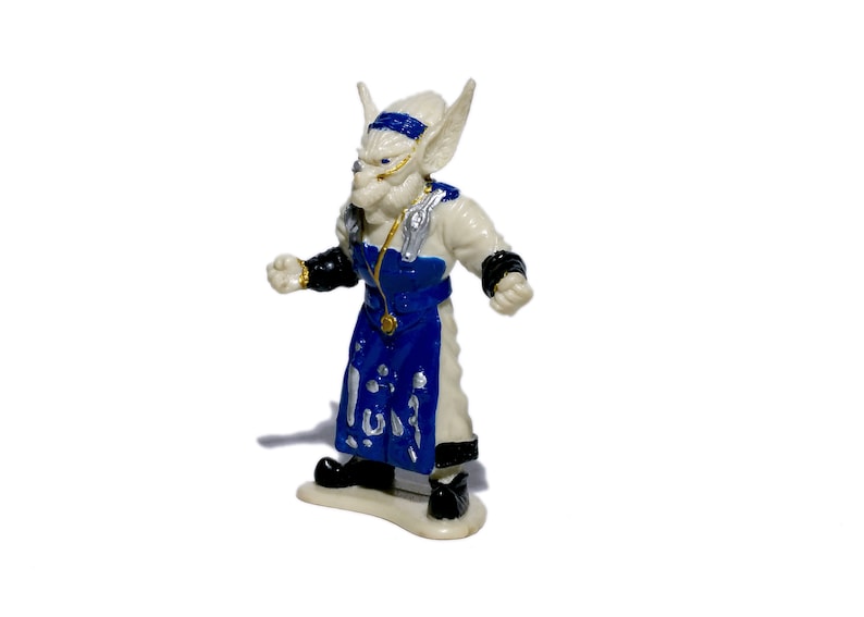 Puede incluir: Una figurita de pl&aacute;stico blanca de una criatura fant&aacute;stica con detalles azules y dorados. La criatura tiene orejas grandes, una nariz puntiaguda y lleva una t&uacute;nica azul y blanca.