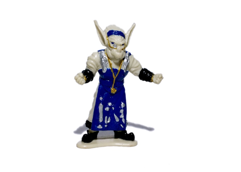 Puede incluir: Una figurita de pl&aacute;stico blanco de un goblin que lleva un delantal azul y blanco y una cadena dorada. El goblin tiene orejas grandes y una cara blanca con ojos azules.