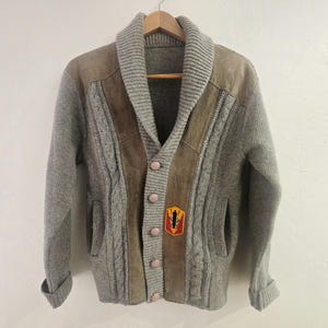 May include: A gray cardigan sweater with a shawl collar and leather accents. The sweater has a cable knit design on the front and sleeves, with a small patch near the bottom. The sweater is buttoned up and hanging on a wooden hanger.