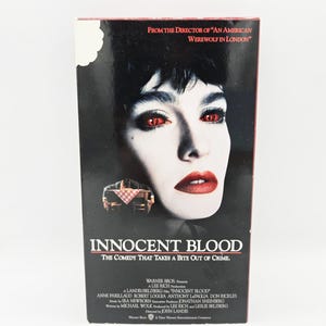 Puede incluir: Una cinta VHS de la película Innocent Blood con un primer plano en blanco y negro de un rostro con ojos y labios rojos. La portada incluye el texto From the director of An American Werewolf in London, el título y los créditos de producción.