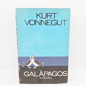 Może przedstawiać: Książka w miękkiej okładce zatytułowana "Galápagos: A Novel" autorstwa Kurta Vonneguta. Okładka przedstawia niebieskie niebo, ciemnoniebieski pas i dwa ptaki. Imię autora jest białe, a tytuł biało-czarny.