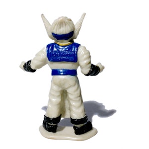 Puede incluir: Una figura de acci&oacute;n de pl&aacute;stico blanco con detalles azules y dorados. La figura tiene orejas puntiagudas y lleva un mono blanco con un cintur&oacute;n azul y un chaleco azul. La figura est&aacute; de pie sobre una base blanca.