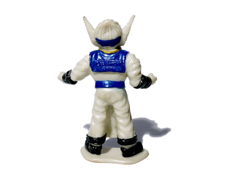 Puede incluir: Una figura de acci&oacute;n de pl&aacute;stico blanco con detalles azules y dorados. La figura tiene orejas puntiagudas y lleva un mono blanco con un cintur&oacute;n azul y un chaleco azul. La figura est&aacute; de pie sobre una base blanca.