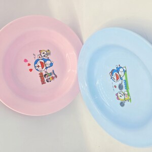 Puede incluir: Dos platos de pl&aacute;stico, uno rosa y otro azul claro, cada uno con un dibujo animado de un personaje robot azul con forma de gato. El plato rosa tiene un dise&ntilde;o de coraz&oacute;n. Los platos son redondos con un borde ligeramente elevado.