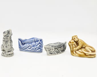 May include: Four small ceramic figurines. A gray wolf, a blue boat with a fish, a gray boat, and a tan monkey resting on a tree stump. The figurines are on a white surface.